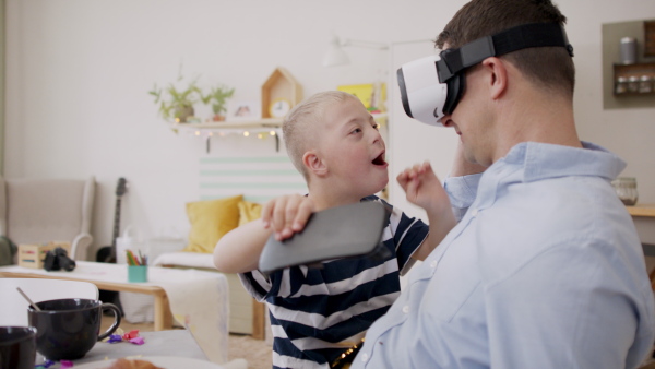 A father with happy down syndrome son indoors at home, using vr goggles.