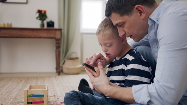 A father with happy down syndrome son indoors at home, using smartphone.