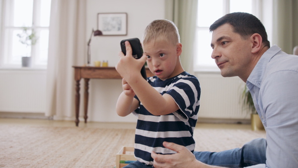 A father with happy down syndrome son indoors at home, using smartphone.