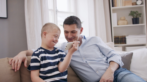 A father with happy down syndrome son indoors at home, using smartphone.