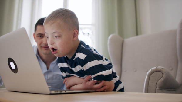 A father with happy down syndrome son indoors at home, using laptop.