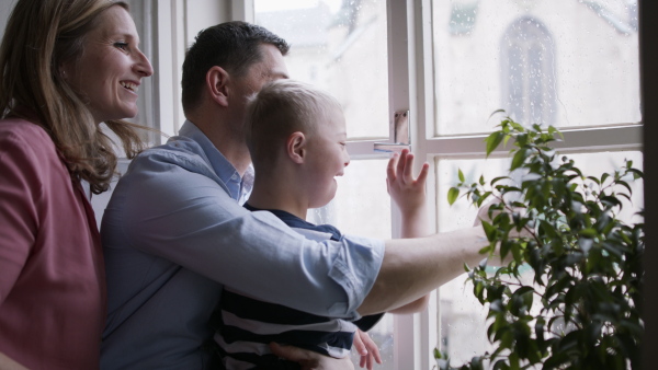 A family with happy down syndrome son indoors at home, looking through window and waving.