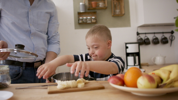 A father with happy down syndrome son indoors in kitchen, cooking.