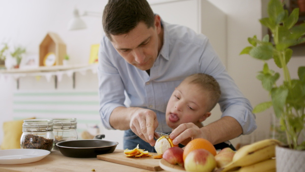 A father with happy down syndrome son indoors in kitchen, preparing food.