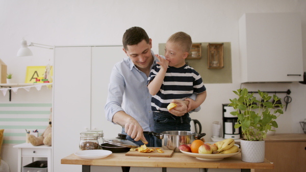 A father with happy down syndrome son indoors in kitchen, preparing food.