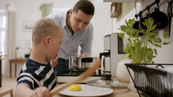 A father with happy down syndrome son indoors in kitchen, cooking.