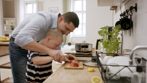 A father with happy down syndrome son indoors in kitchen, chopping apple.