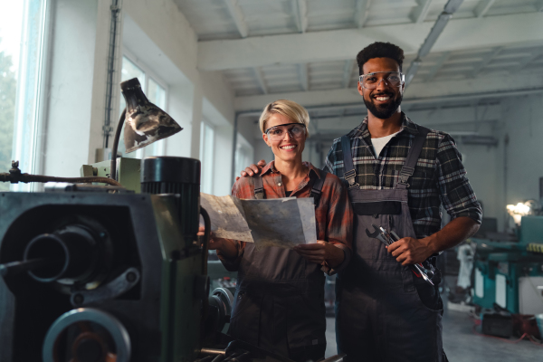 A portrait of young biracial industrial colleagues working indoors in metal workshop, smiling and looking at camera.