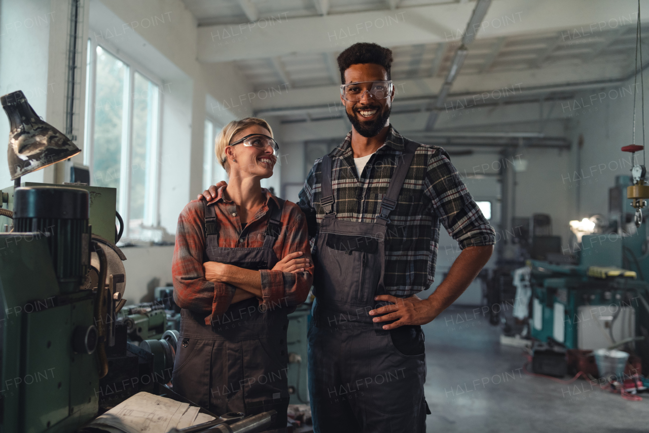 A portrait of young biracial industrial colleagues working indoors in metal workshop, smiling and looking at camera.