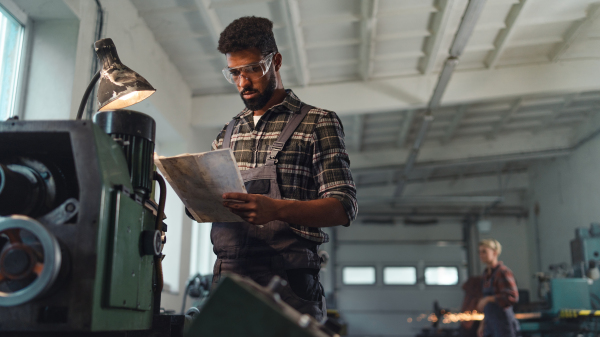 A low angle view of young industrial man working indoors in metal workshop.