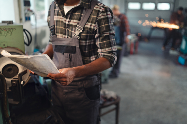 A close up of young industrial man holding blueprints indoors in metal workshop.