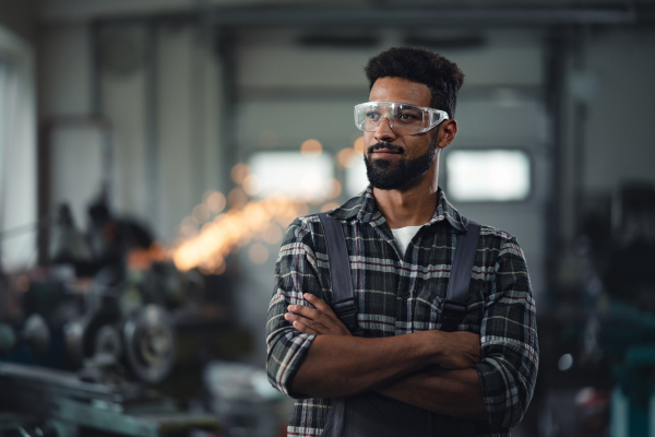 A portrait of young industrial man standing with arms crossed indoors in metal workshop, looking away.