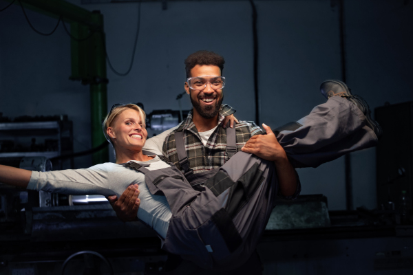 A portrait of young African American man lifting up his female colleague indoors in metal workshop.