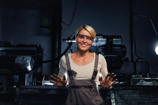 Portrait of a happy young industrial woman with dirty hands looking at camera indoors in metal workshop.