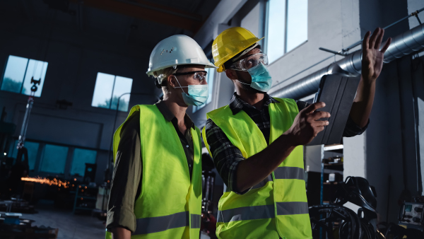 A low angle view of industrial inspectors with face masks doing a general check up indoors at metal workshop, coronavirus concept.