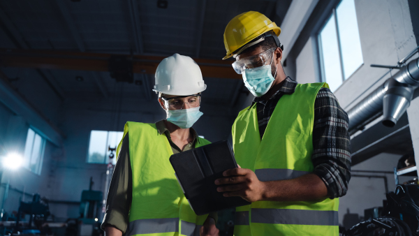 A low angle view of industrial inspectors with face masks doing a general check up indoors at metal workshop, coronavirus concept.