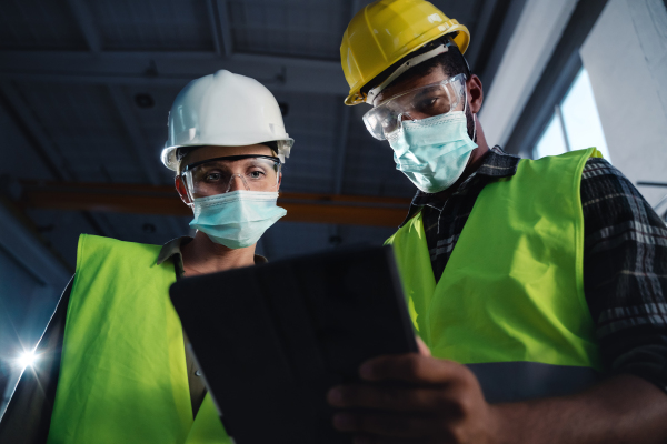 A low angle view of industrial inspectors with face masks doing a general check up indoors at metal workshop, coronavirus concept.