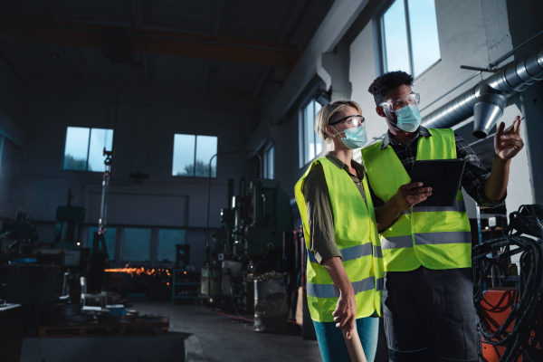 An industrial inspectors with face masks doing a general check up indoors at metal workshop, coronavirus concept.