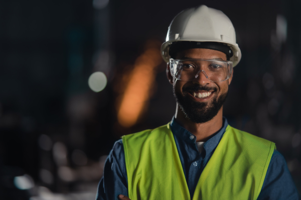 A portrait of happy young industrial man working indoors in metal workshop, looking at camera.