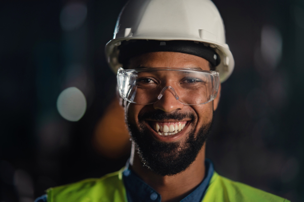 A close up portrait of happy young industrial man with protective wear indoors in metal workshop, looking at camera.