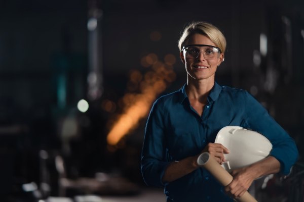 A portrait of happy young industrial woman engineer looking at camera indoors in metal workshop.