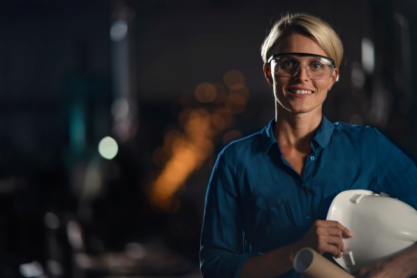 A portrait of mid adult industrial woman working indoors in metal workshop, looking at camera.