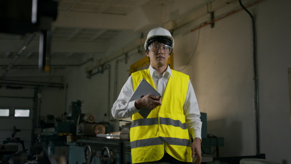 A young industrial Japanese man engineer with protective wear using tablet indoors in metal workshop.