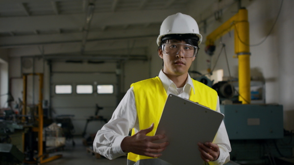 A young industrial Japanese man engineer with protective wear indoors in metal workshop, looking at camera.