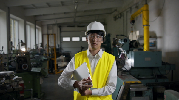 A young industrial Japanese man engineer with check list indoors in metal workshop, looking at camera.