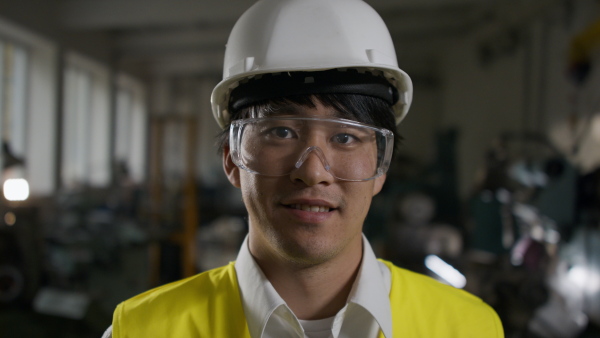 A close up of young industrial Japanese man engineer looking at camera indoors in metal workshop.