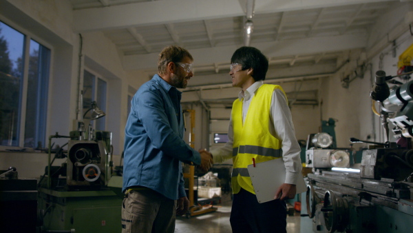 Japanese engineer shaking hands with a mature wroker indoors in metal workshop.