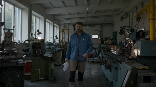 A mature industrial man working indoors in metal workshop, looking at camera.