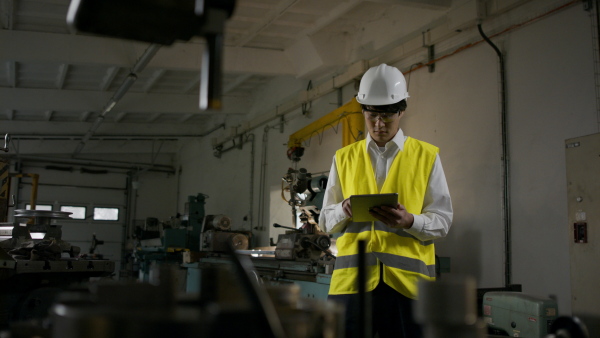 A young industrial Japanese man engineer with protective wear using tablet indoors in metal workshop.