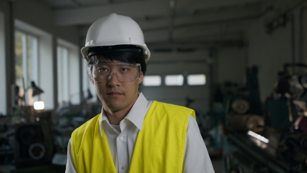 A young industrial Japanese man engineer with protective wear indoors in metal workshop, looking at camera.