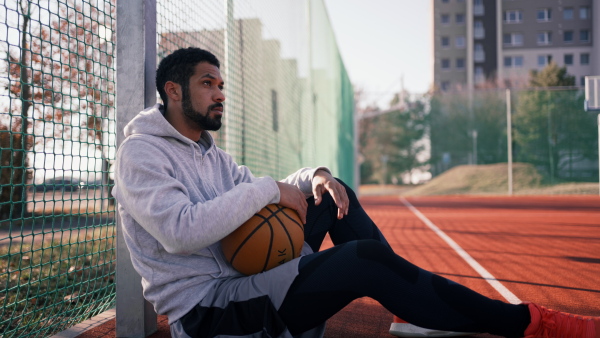 Young black Man sitting on basketball playground, holding a ball, resting.