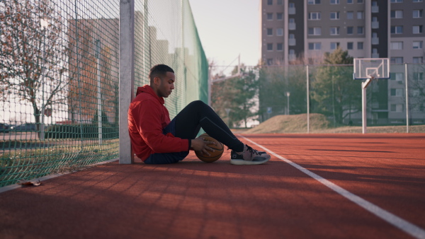 Young pensive african american man at urban playground in sports clothes playing basketball, resting and sitting at basketball court.