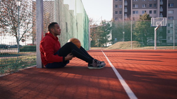 Young black basketball player sitting on basketball playground, holding a ball, looking into camera, smiling.