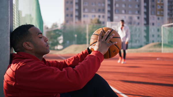 Close up of young american african basketball player, sitting on playground, holding the ball, greeting his fellow player.