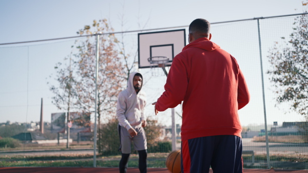 Cheerful friends playing basketball outside, one throwing ball into basket. Back view.