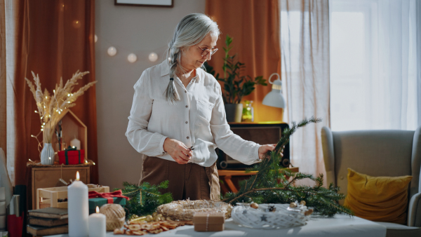 Senior woman cutting a branch to make Christmas wreath indoors at home