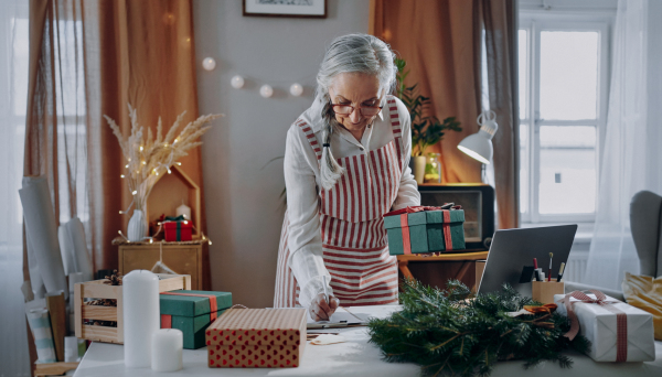 A senior woman packing Christmas presents and writing notes to list indoors, small business concept.