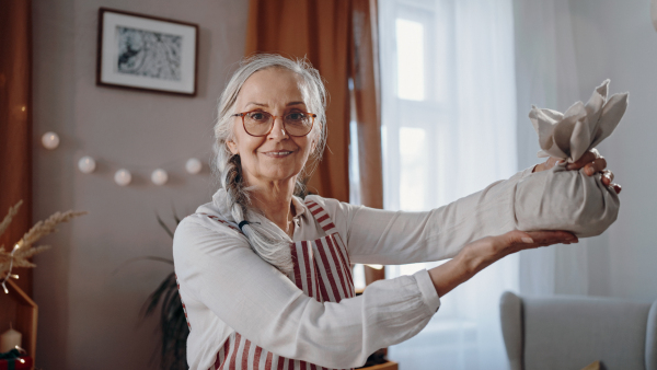 A senior woman holding Christmas present wrapped in eco materials indoors at home, looking at camera