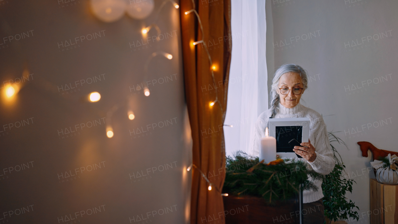 A nostalgic senior woman looking at picture frame, standing by window at home.