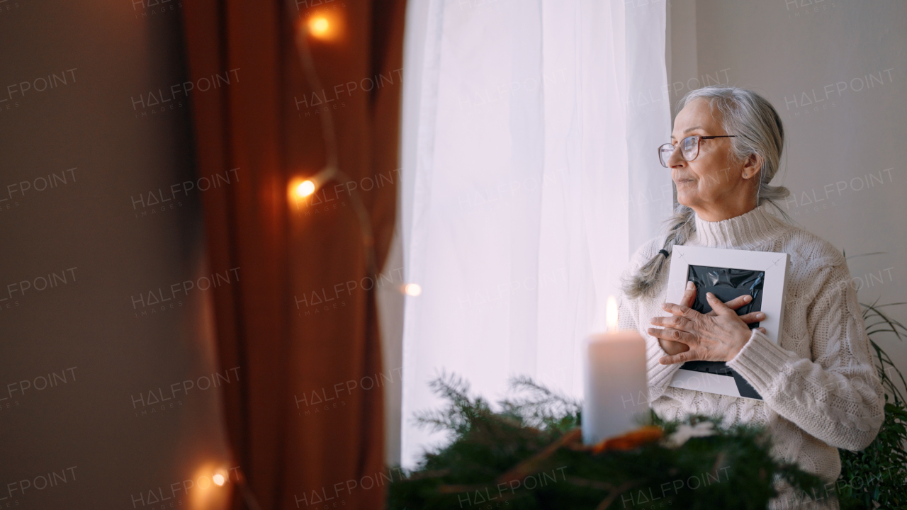 A nostalgic senior woman holding picture frame, standing by window at home.