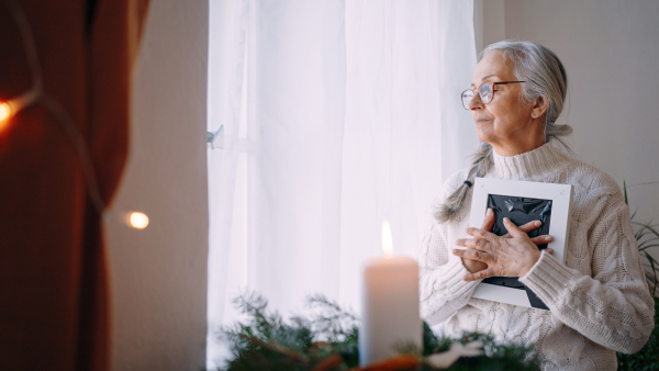 A sad senior woman with eyes closed hugging picture frame, standing by window at home.