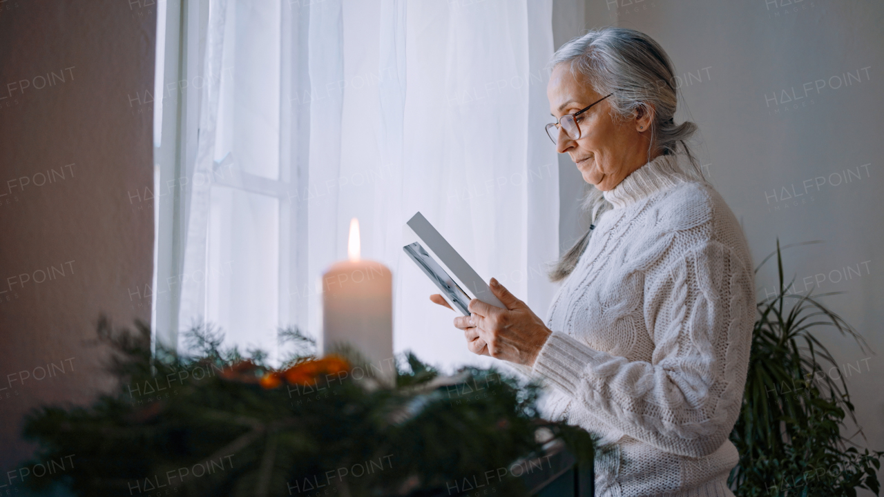 A nostalgic senior woman looking at picture frame, standing by window at home.