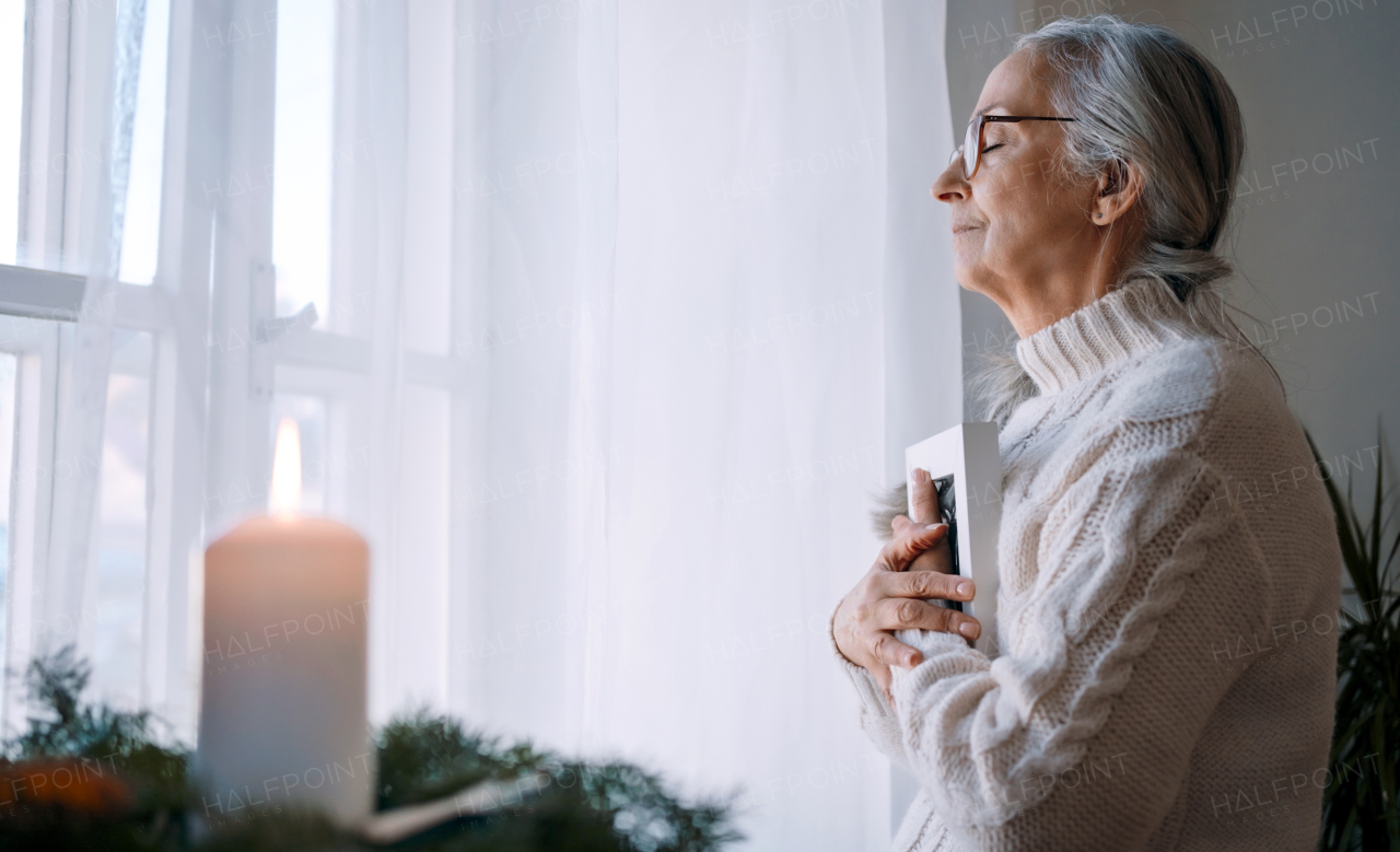 A sad senior woman with eyes closed hugging picture frame, standing by window at home.