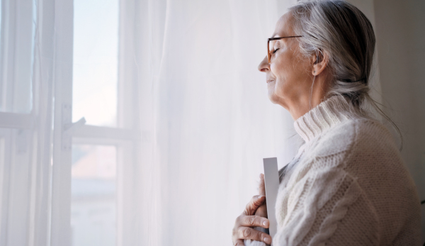 A sad senior woman with eyes closed standing by window and holidng a picture frame indoors at home