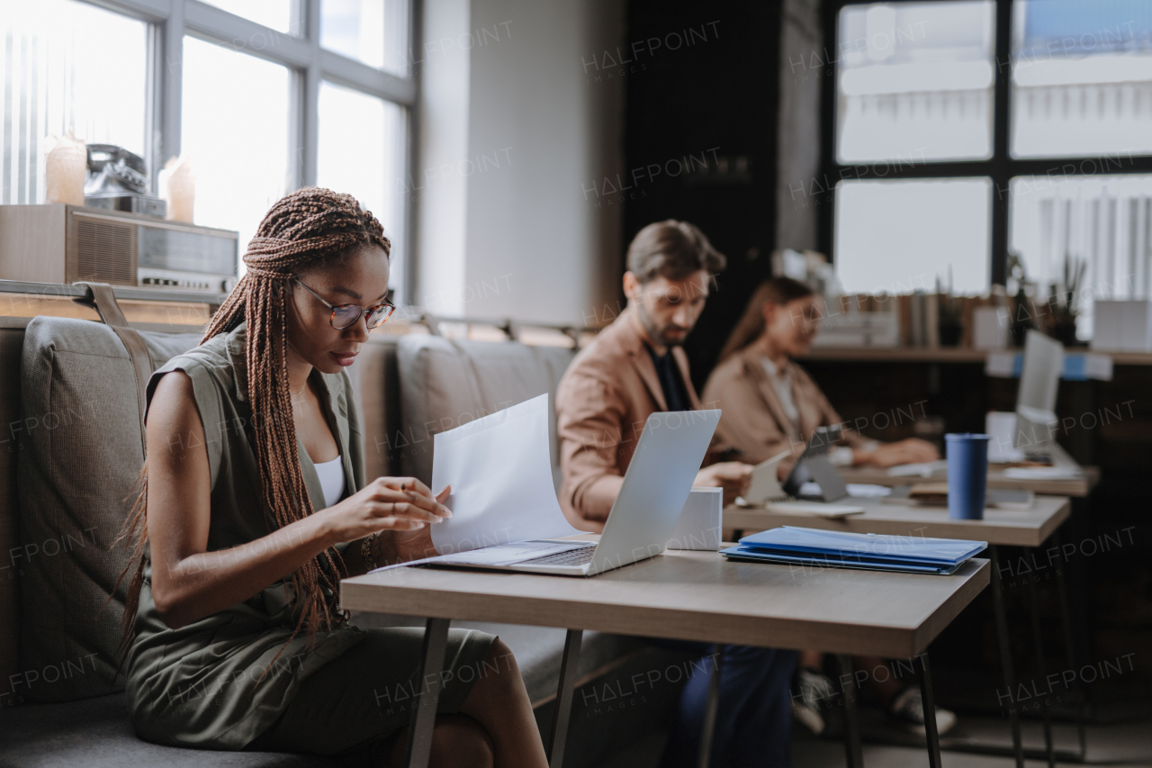 Beautiful businesswoman working on laptop, reading document in modern office. Group of freelancers working in shared work area. Concept of coworking, common workspaces for business, startups. Working remotely from coffee shop.