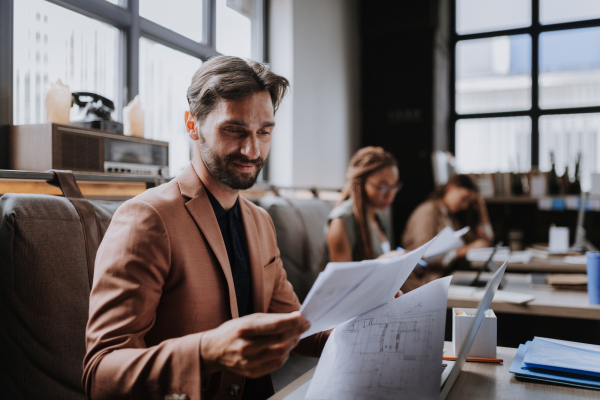 Businessman working on laptop and reading documents in modern office or coworking area. Working remotely from the coffee shop. Concept of coworking, common workspaces for business, startups.